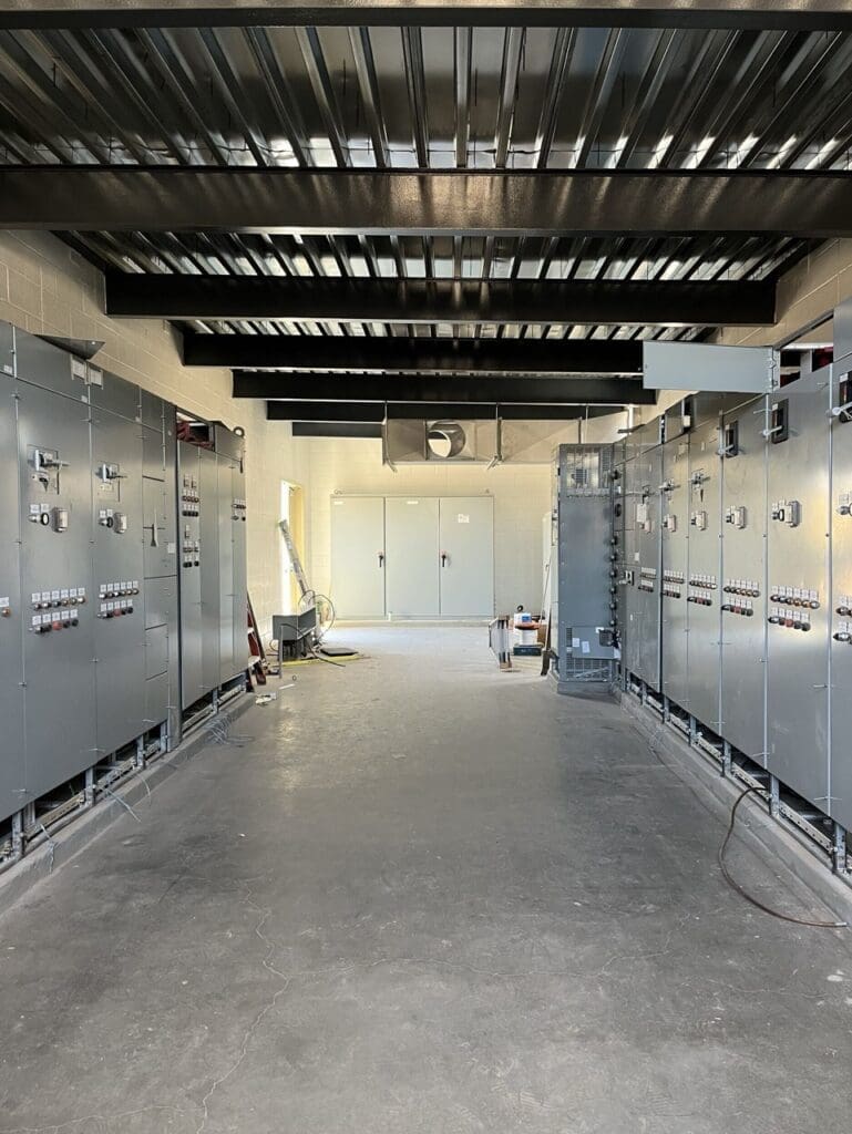 Electrical control room with installed switchgear assemblies, overhead ventilation ductwork, and exposed steel roof framing during final installation phase at City of Phoenix Booster Pump Station