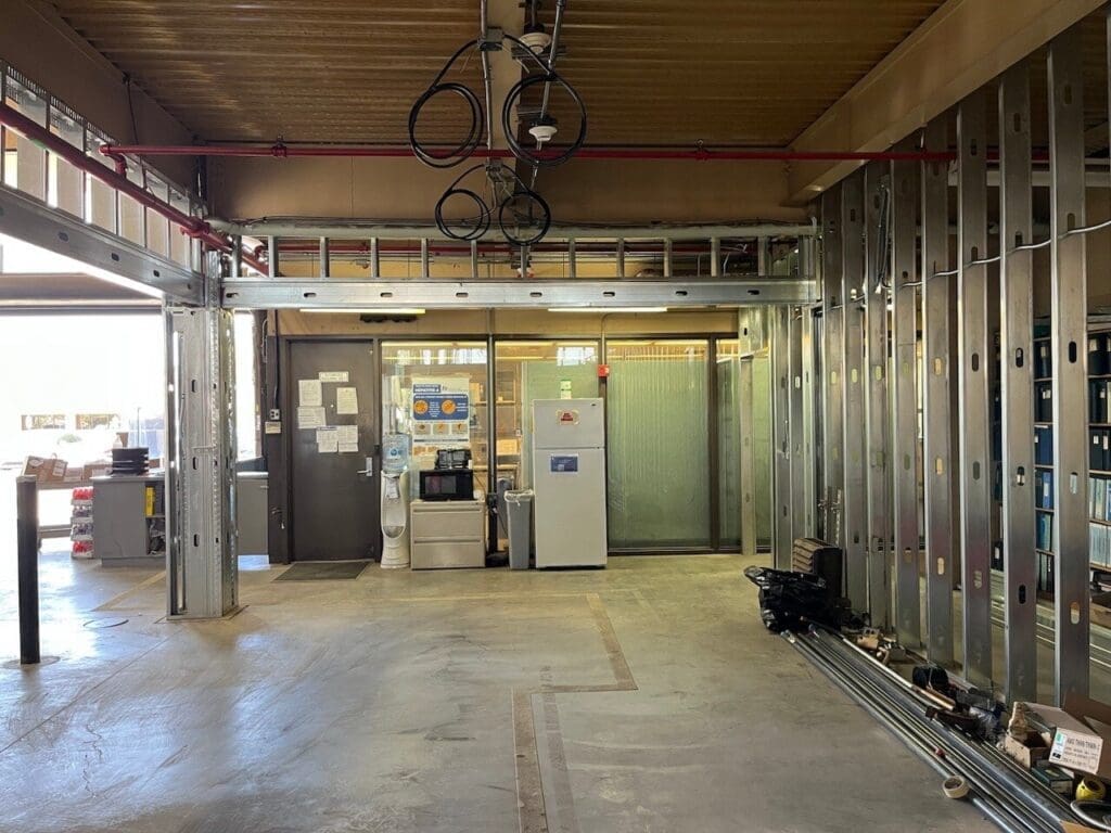 Interior warehouse space with steel stud framing for office build-out, exposed ceiling utilities, and break area with refrigerator and water cooler