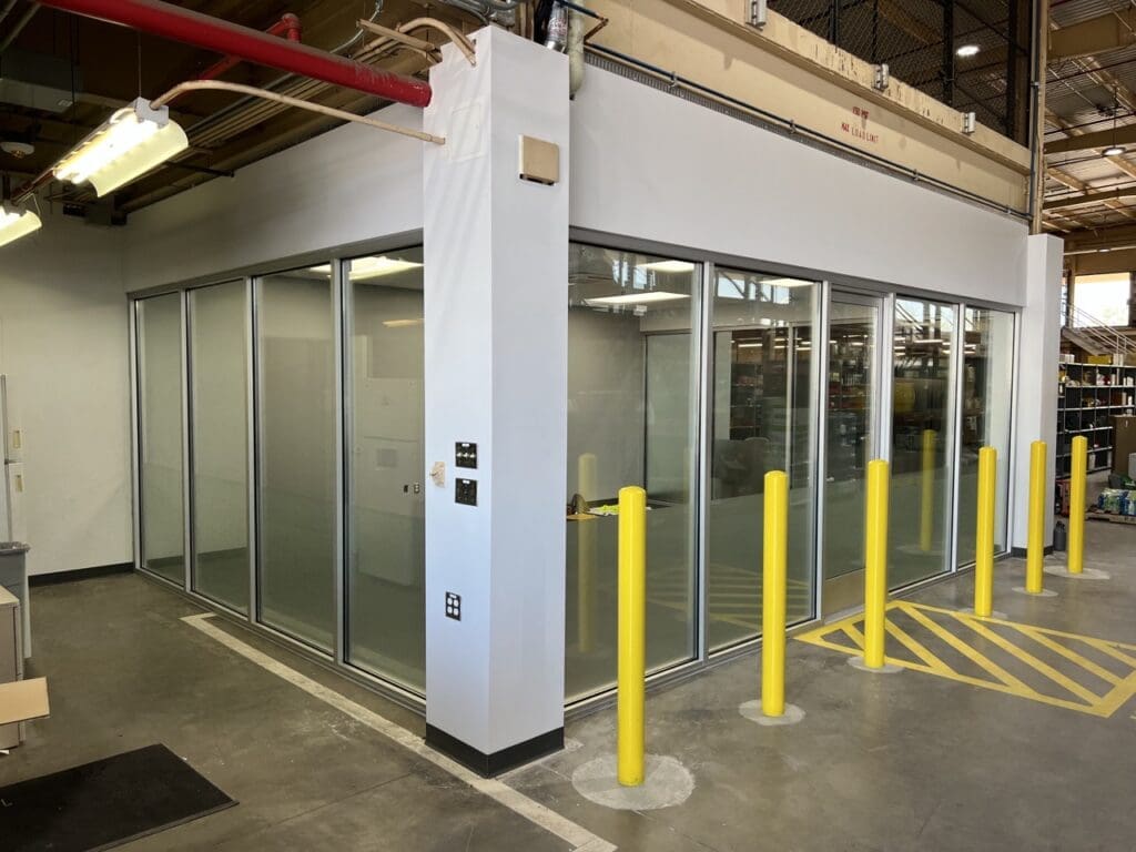 Finished interior office enclosure with glass walls inside warehouse, featuring safety bollards and overhead utility piping.