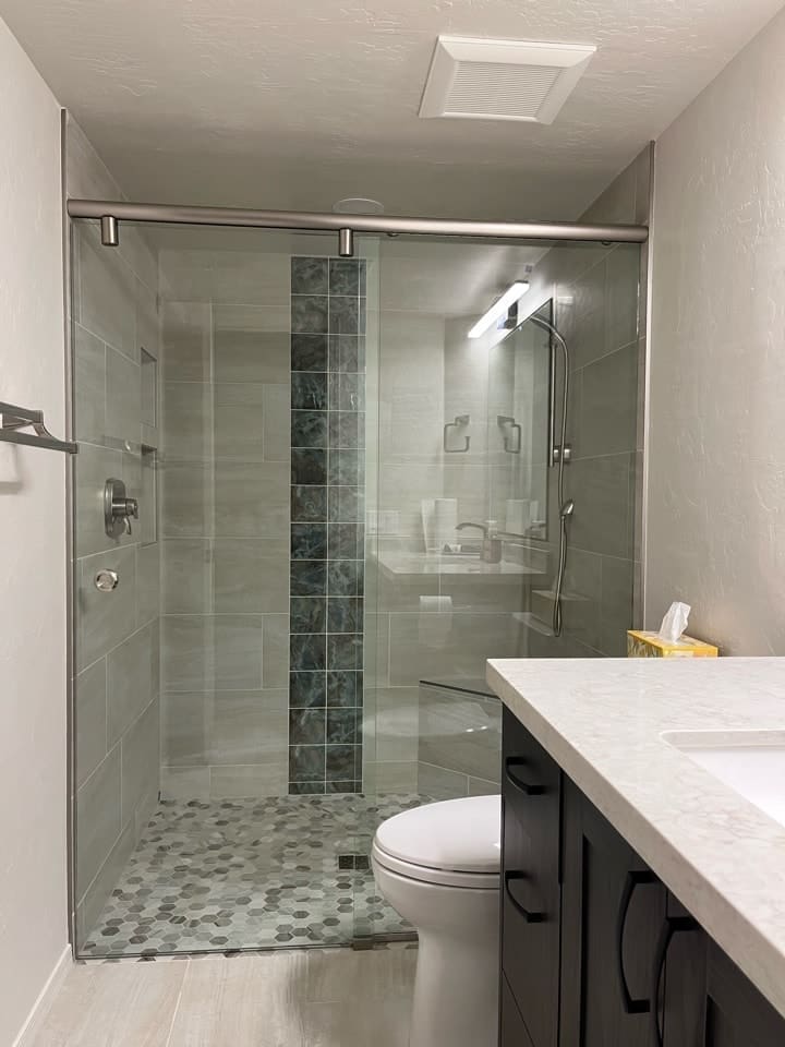 Contemporary bathroom remodel featuring a frameless glass walk-in shower, custom tile walls with decorative accent strip, pebble-style shower flooring, quartz vanity countertop, matte black cabinetry hardware, and upgraded modern fixtures for a clean spa-inspired finish.