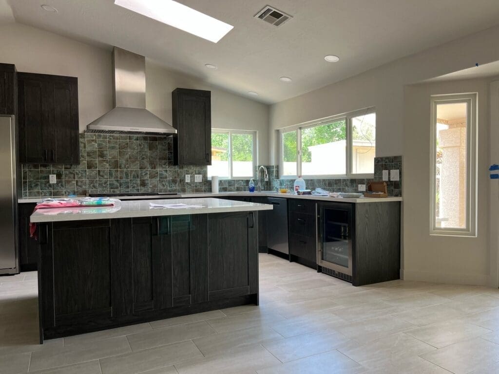 Completed Ahwatukee kitchen remodel featuring a large quartz waterfall island, custom dark wood cabinetry, stainless steel range hood, expansive window lighting, modern tile backsplash, skylight feature, and upgraded open-concept kitchen finishes with contemporary luxury styling.