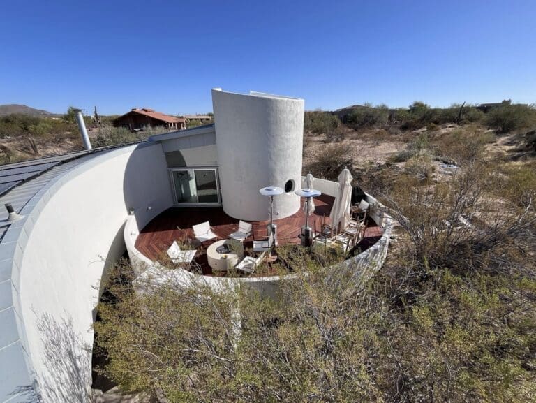 Completed curved outdoor deck and patio area in a desert-style modern home, featuring custom geometric wood decking, built-in seating walls, lounge furniture, patio heaters, and a minimalist stucco exterior integrated into a natural arid landscape.