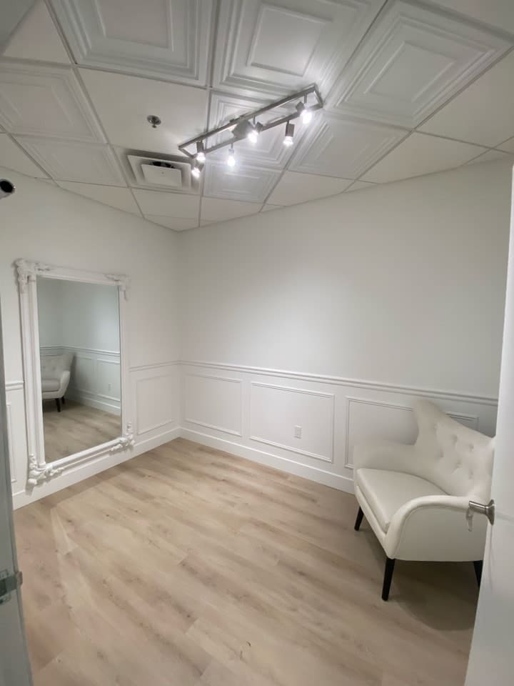 Elegant fitting room with light wood flooring and white paneled walls, featuring a large decorative full-length mirror with an ornate frame, overhead track lighting, and a white upholstered accent chair positioned in the corner.