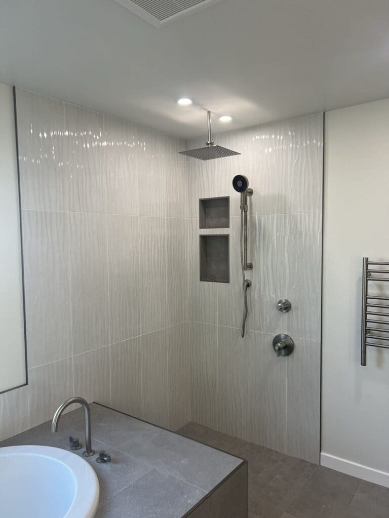 Modern walk-in shower with large-format textured tile, rainfall showerhead, built-in niches, and frameless glass enclosure beside soaking tub.