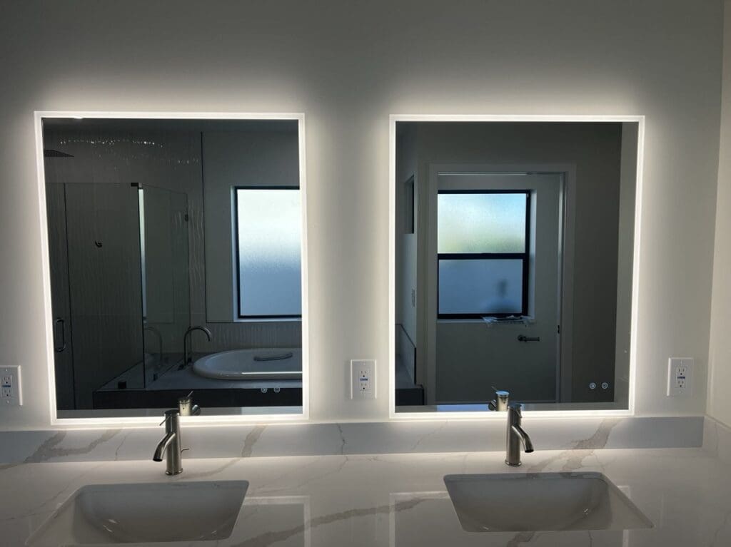 Modern double vanity with quartz countertop, under-mount sinks, and backlit mirrors, reflecting a glass-enclosed shower and soaking tub.
