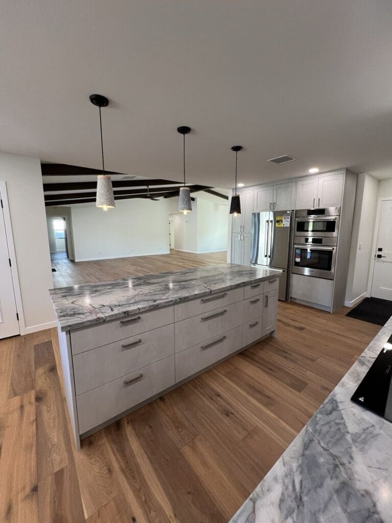 Spacious modern kitchen with a large marble waterfall island, pendant lighting, stainless steel appliances, and warm wood flooring in an open-concept layout.