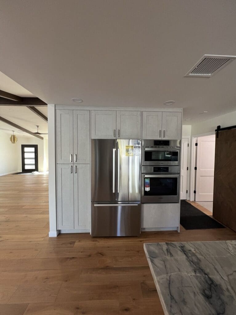 Modern kitchen wall with integrated stainless steel refrigerator and double oven, framed by light wood cabinetry and set against warm wood flooring in an open-concept space.