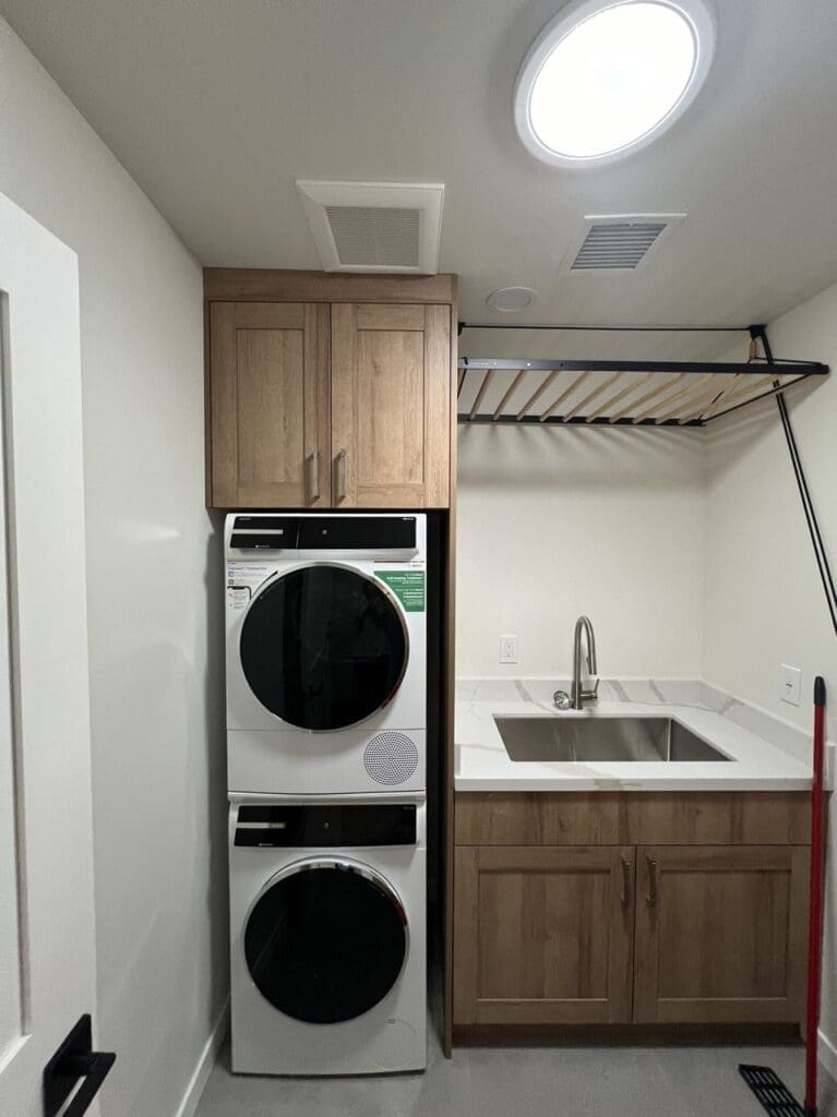 Compact laundry room with stacked washer and dryer, warm wood cabinetry, a utility sink with quartz countertop, and overhead drying rack for added functionality.