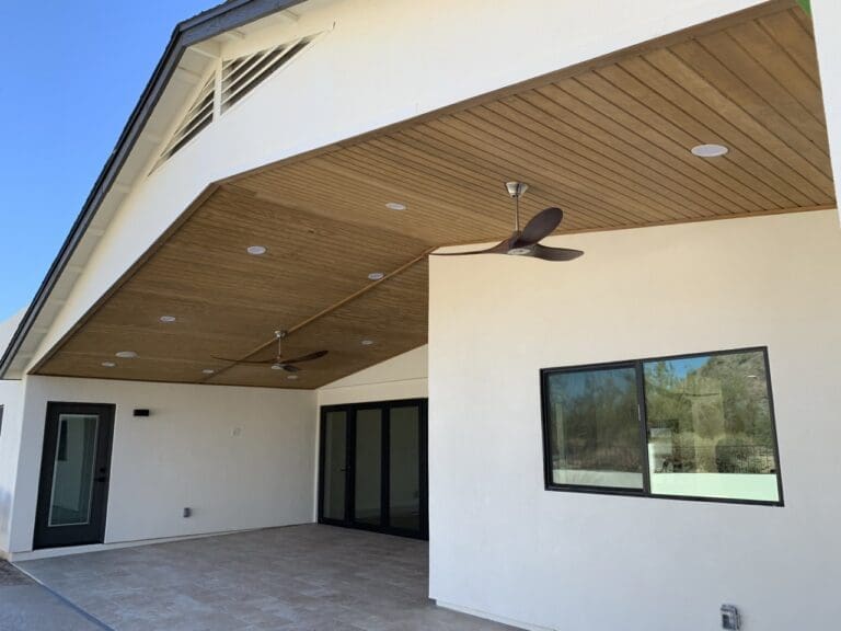 Covered patio with wood-paneled ceiling, recessed lighting, and dual ceiling fans, opening to a modern outdoor living space with sliding glass doors.