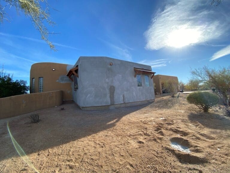Exterior view of a newly stuccoed home addition with desert landscaping and bright sunlight in the background