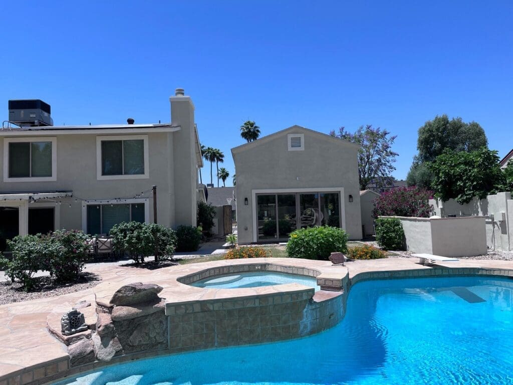 Backyard view of a modern detached casita conversion overlooking a resort-style pool, featuring contemporary stucco architecture, large glass doors, and landscaped outdoor living space.