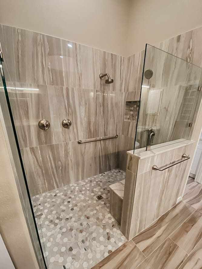 Spacious walk-in shower featuring large striped wall tiles, hexagon floor tile, dual controls, a built-in bench, and multiple grab bars