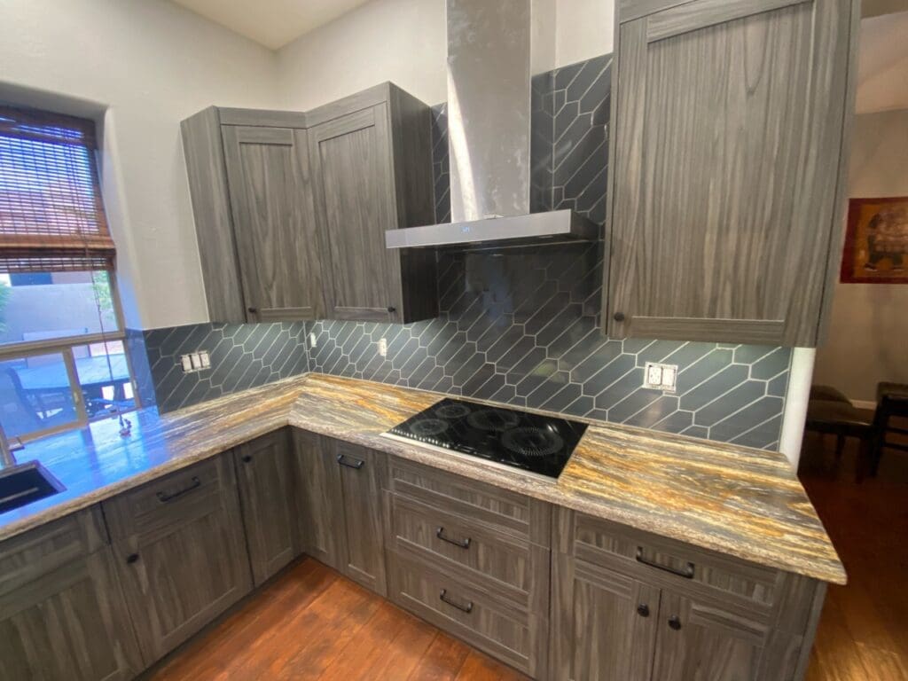Modern cooking area featuring textured gray cabinetry, dramatic stone countertops, a sleek black geometric tile backsplash, and a stainless steel range hood for a bold contemporary finish.