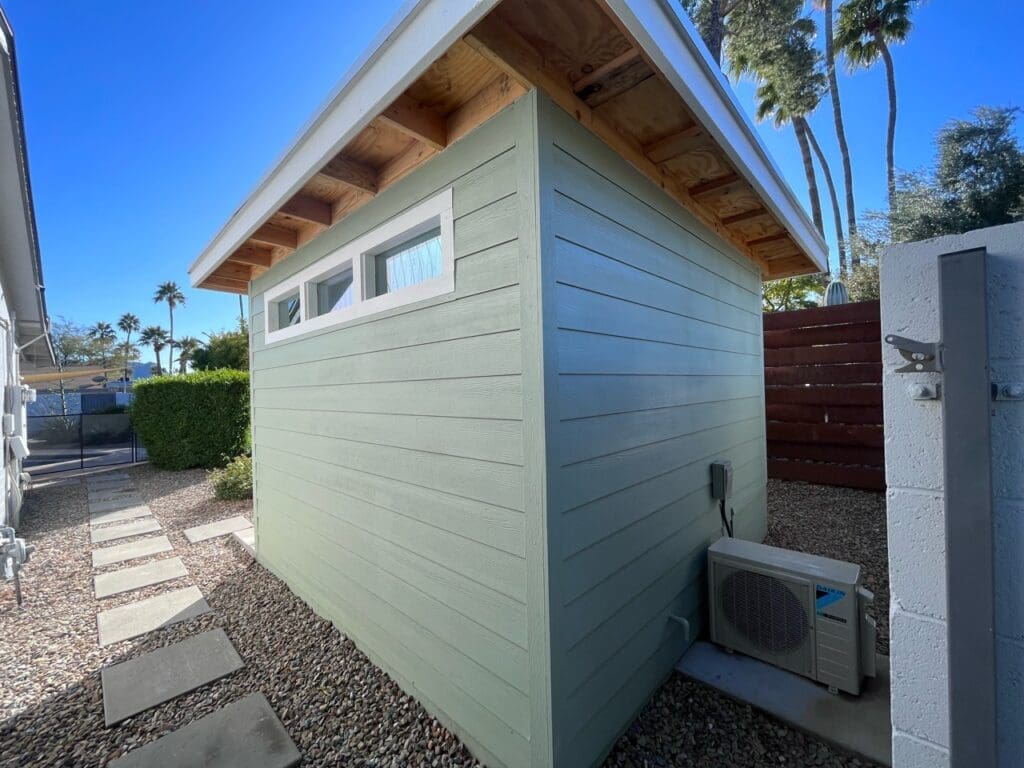 Side view of a small green shed with horizontal siding, clerestory windows, and an exterior HVAC unit set along a gravel pathway
