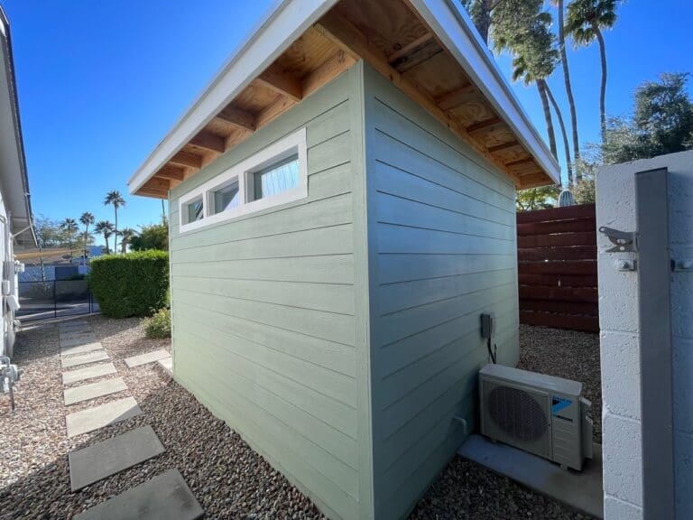 Side view of a small green shed with horizontal siding, clerestory windows, and an exterior HVAC unit set along a gravel pathway