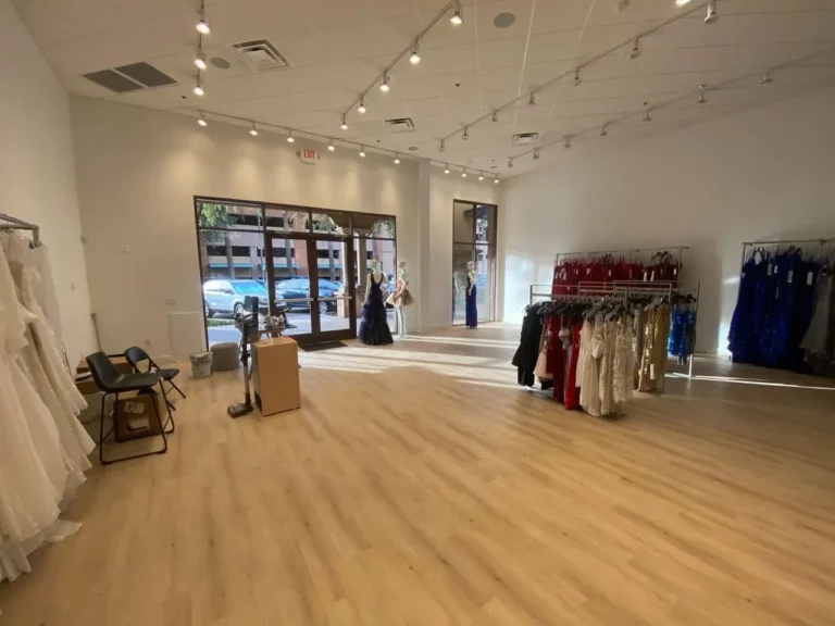 Spacious boutique interior with wooden floors, racks of formal dresses, mannequins near large front windows, and track lighting across the ceiling