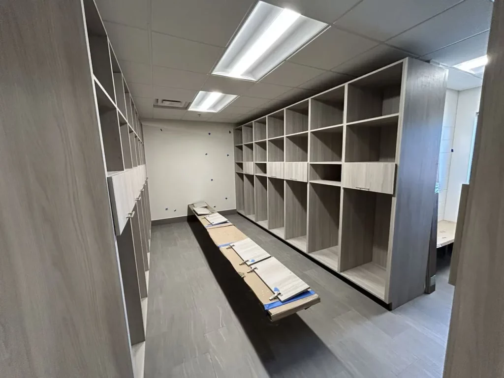 Locker room or storage area under construction with built-in wooden cubbies and a central bench, unfinished walls, and overhead lighting