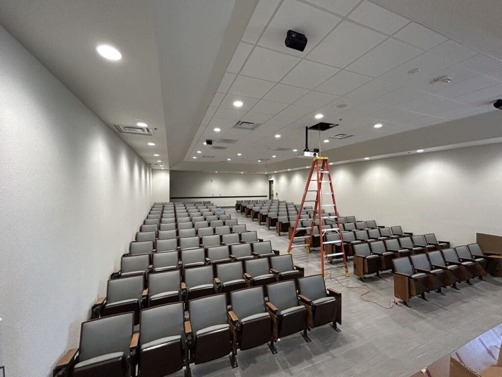 Empty auditorium with rows of gray seats and a ladder set up beneath the ceiling, where a projector or fixture is being installed