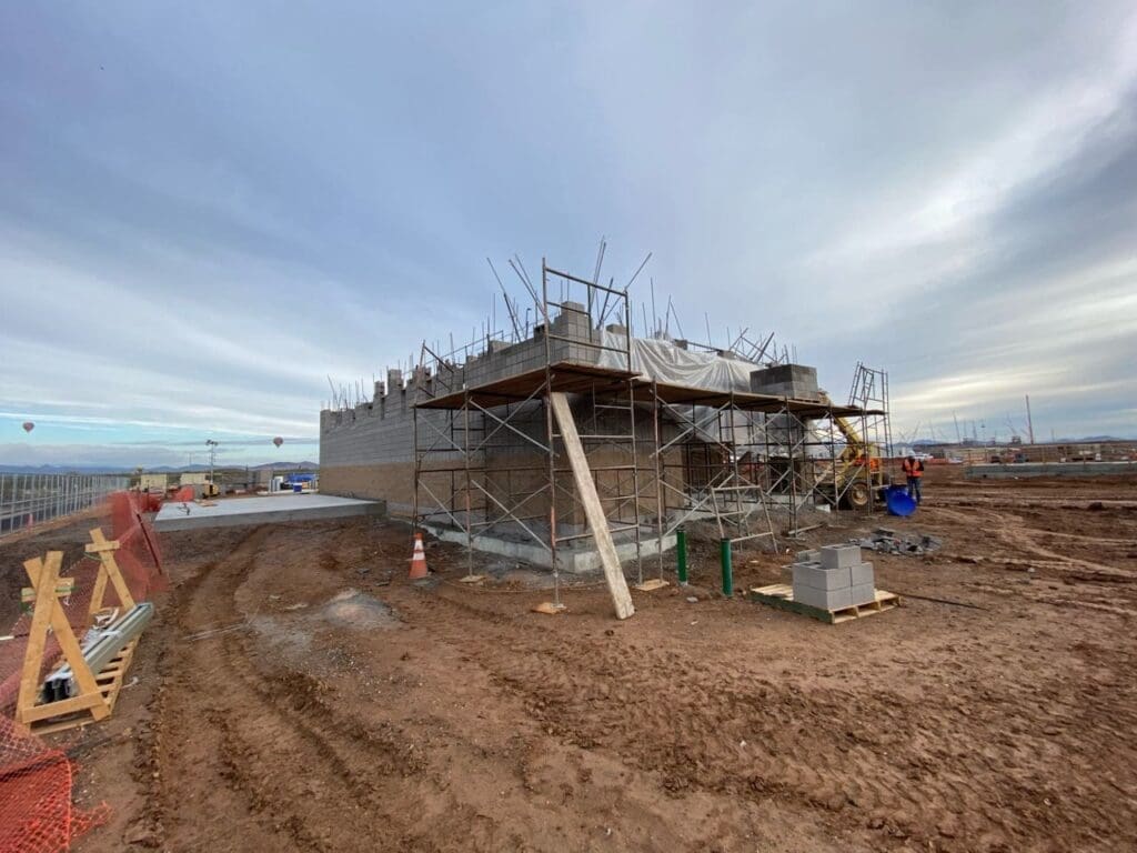 Building under construction with concrete block walls and scaffolding, surrounded by a muddy worksite under a cloudy sky.