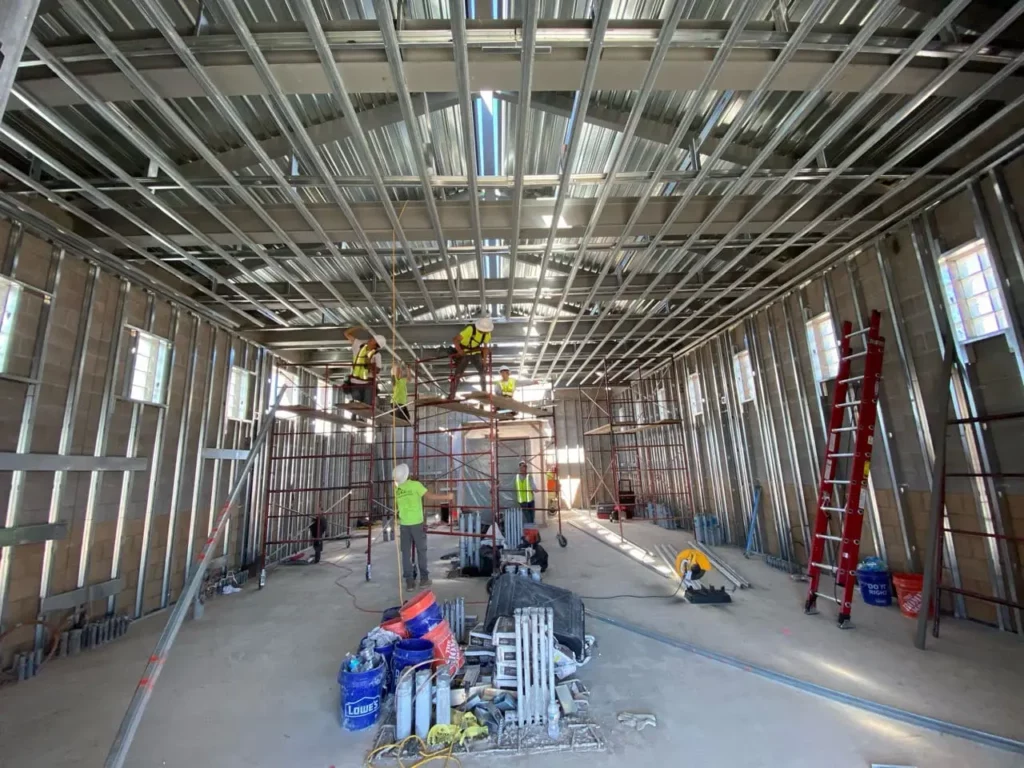 Construction workers installing metal ceiling framing inside a building, using scaffolding and tools amid exposed walls and materials