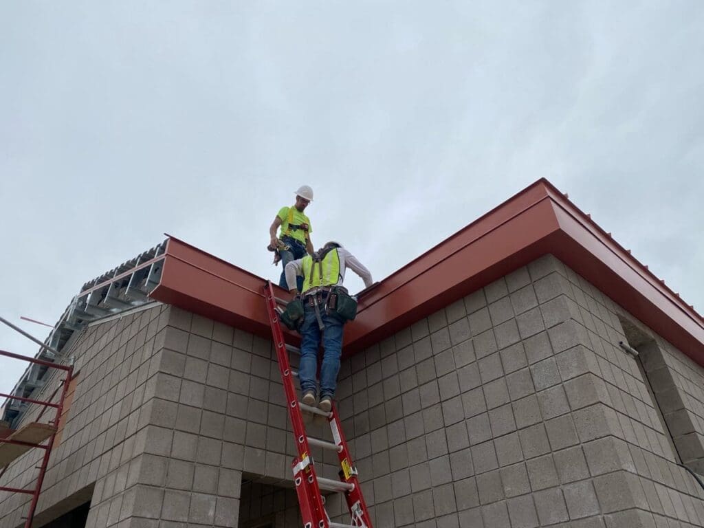 Two construction workers wearing safety gear work on a building roof edge, one standing on a ladder while the other stands above, under an overcast sky