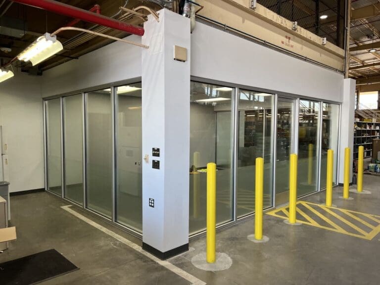 A glass-walled office enclosure inside an industrial warehouse, surrounded by yellow safety bollards and concrete flooring