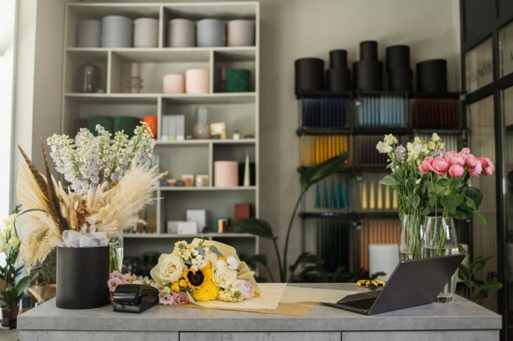 Floral shop workspace with bouquets of flowers on a counter, a laptop, and shelves of vases and supplies in the background