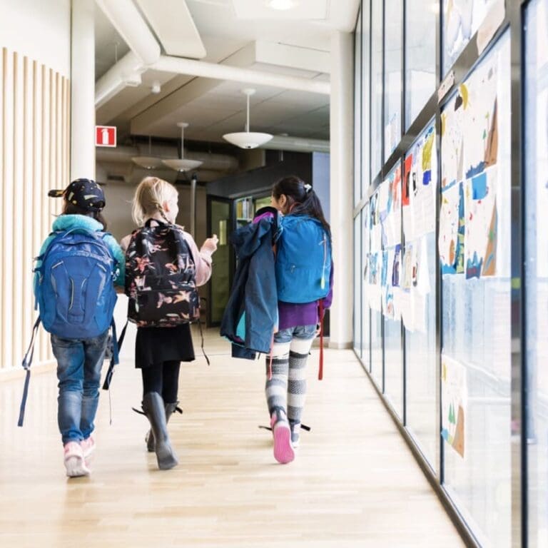 Three children wearing backpacks walk down a school hallway beside a wall displaying colorful artwork