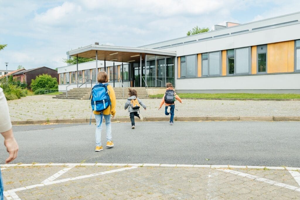 Two children with backpacks run toward a school building while an adult follows behind them in a parking area