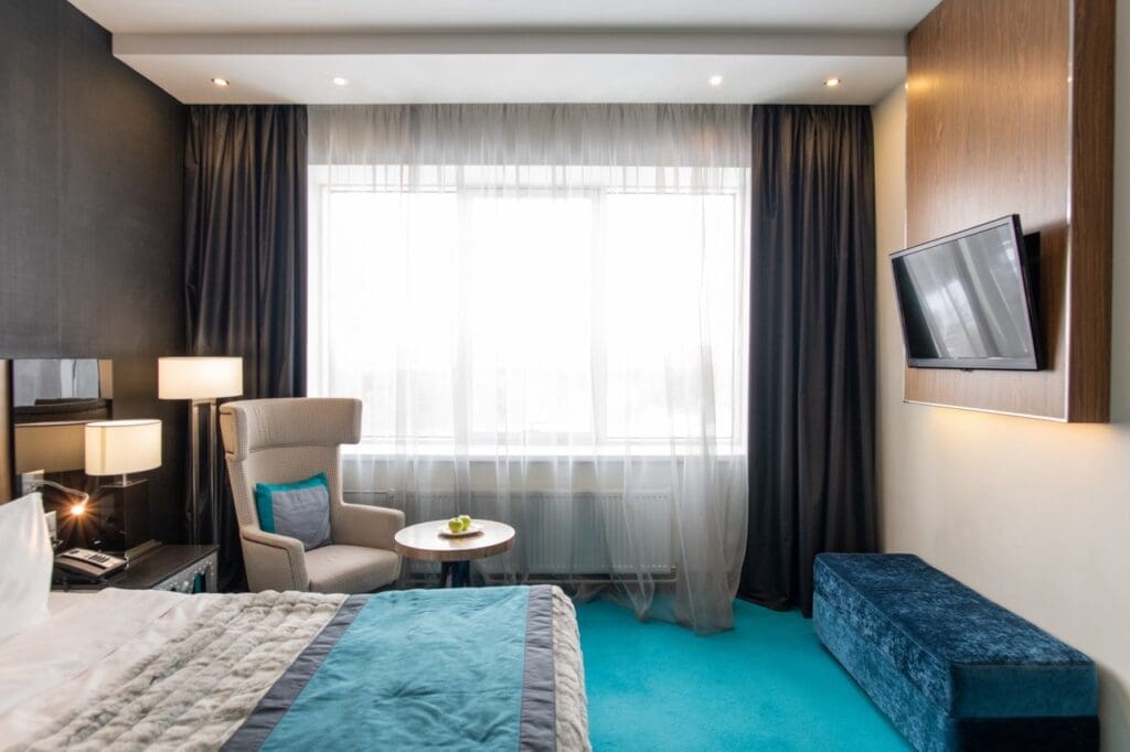 Cozy modern hotel room with bed, armchair, small table, wall-mounted TV, and large window with curtains