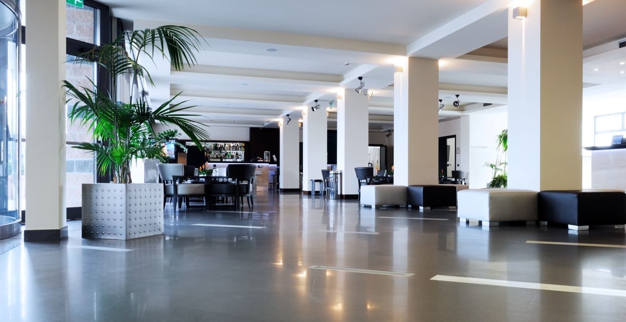 Spacious modern lobby or lounge area with seating, large columns, indoor plants, and a bar or reception area in the background
