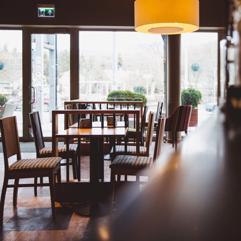 A cozy café interior with wooden tables and chairs, a large hanging lamp, and floor-to-ceiling windows looking out to the street