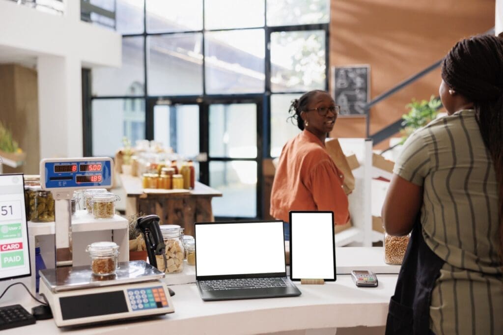 Shop counter with a laptop and tablet on display, as two people interact in a modern grocery or zero-waste store with jars and bulk goods in the background