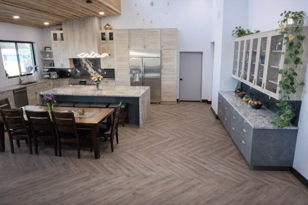 Modern open-concept kitchen and dining area with a large island, stone countertops, light wood cabinetry, stainless steel appliances, and a dining table set nearby, decorated with plants and flowers.