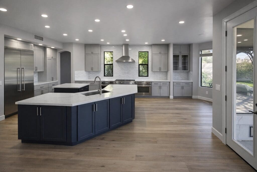 Spacious modern kitchen with a large island, white countertops, gray cabinetry, stainless steel appliances, and wood flooring, brightly lit with recessed ceiling lights