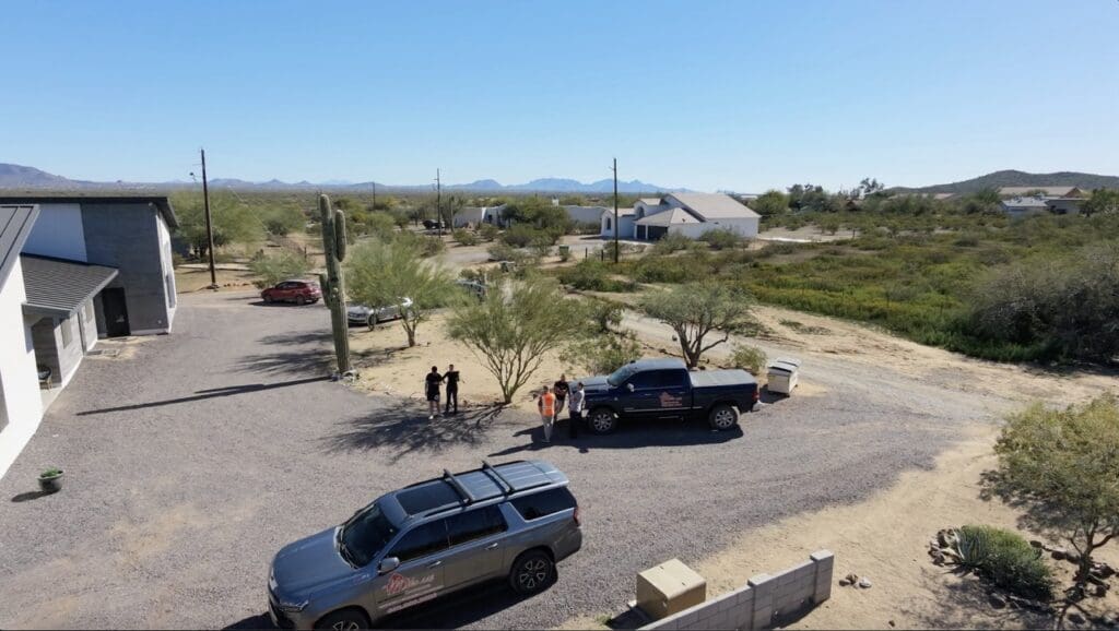 Aerial view of a desert residential area with scattered homes, dirt roads, and sparse vegetation, where several people, including a Miko LLC worker in a high-visibility vest, stand near company vehicles in an open gravel lot